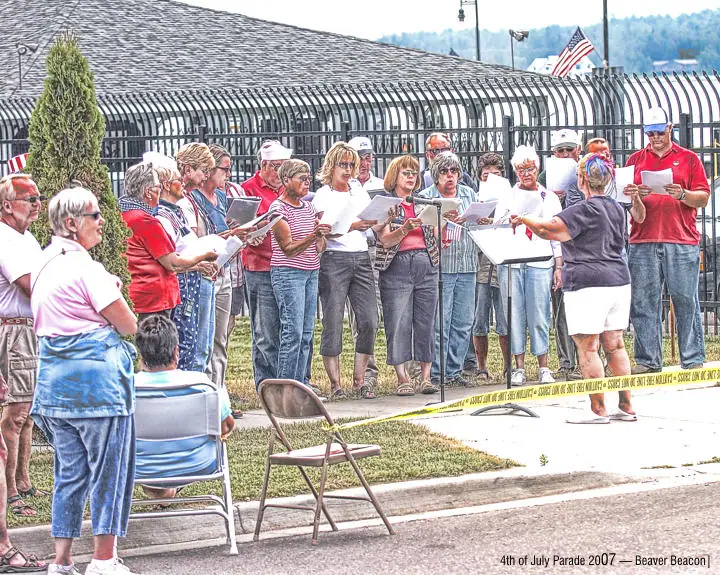 A group sings from printed sheets while a conductor leads them beside a parade route marked with caution tape.