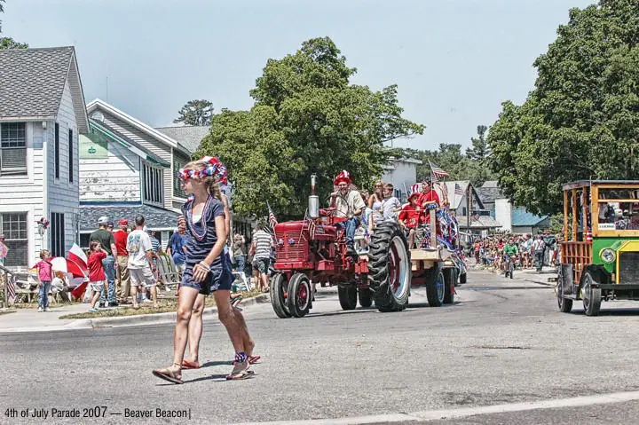 A parade with a decorated tractor, costumed participants, spectators, and a green-and-yellow vehicle on a paved street.