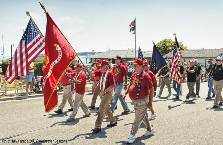 Marchers carrying American and Marine Corps flags proceed along a waterfront street.