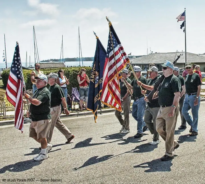Men carrying American and veterans’ flags march past spectators beside sailboats and a waterfront building.