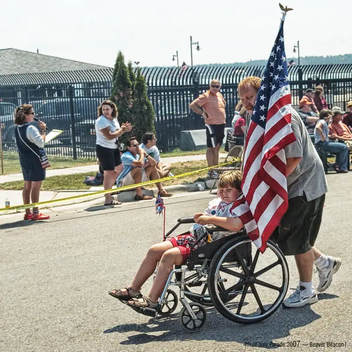 A child in a wheelchair is escorted along a parade route beneath a large American flag.
