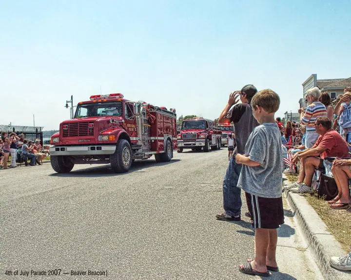 A red fire truck leads a parade past spectators seated along a sunny street.