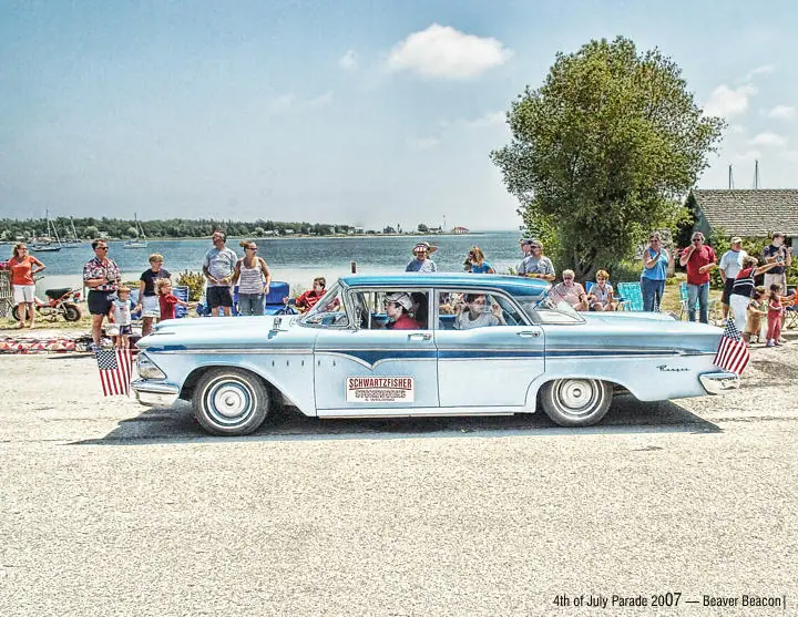 A vintage light-blue sedan decorated with flags passes spectators beside the water during a parade.