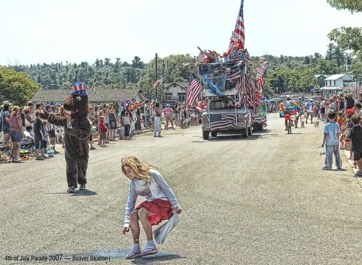 A decorated parade truck follows a bear mascot while a crouching person draws on the pavement.
