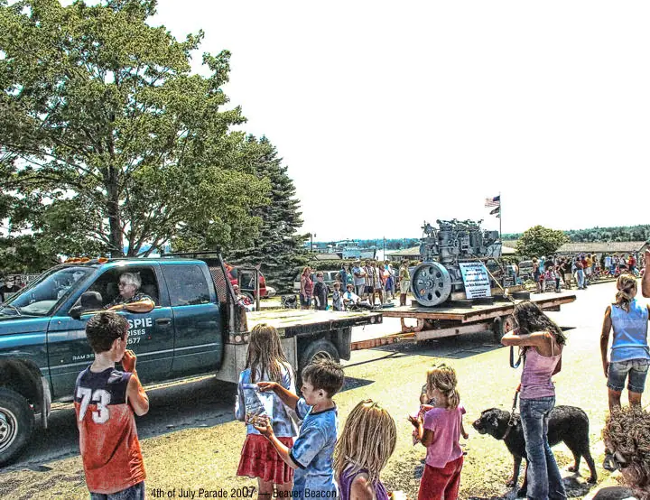 Pickup truck towing a vintage engine past children and spectators during a street parade.