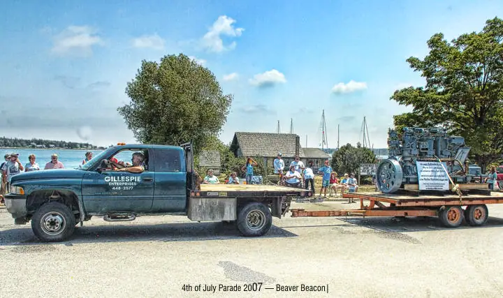 A green truck towing a trailer-mounted industrial engine passes spectators along a waterfront parade route.