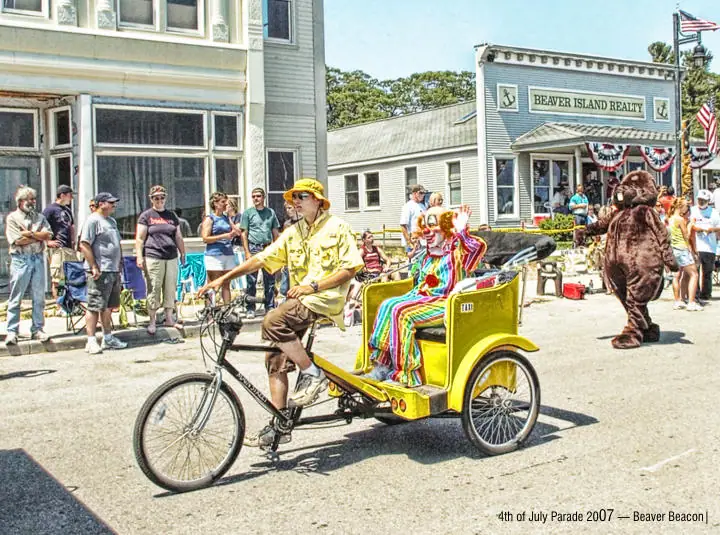 A yellow pedal rickshaw carrying costumed riders passes spectators during a street parade.