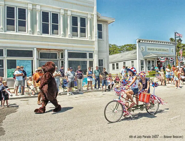 A bear mascot and riders on a decorated tricycle pass a crowd during a summer parade.