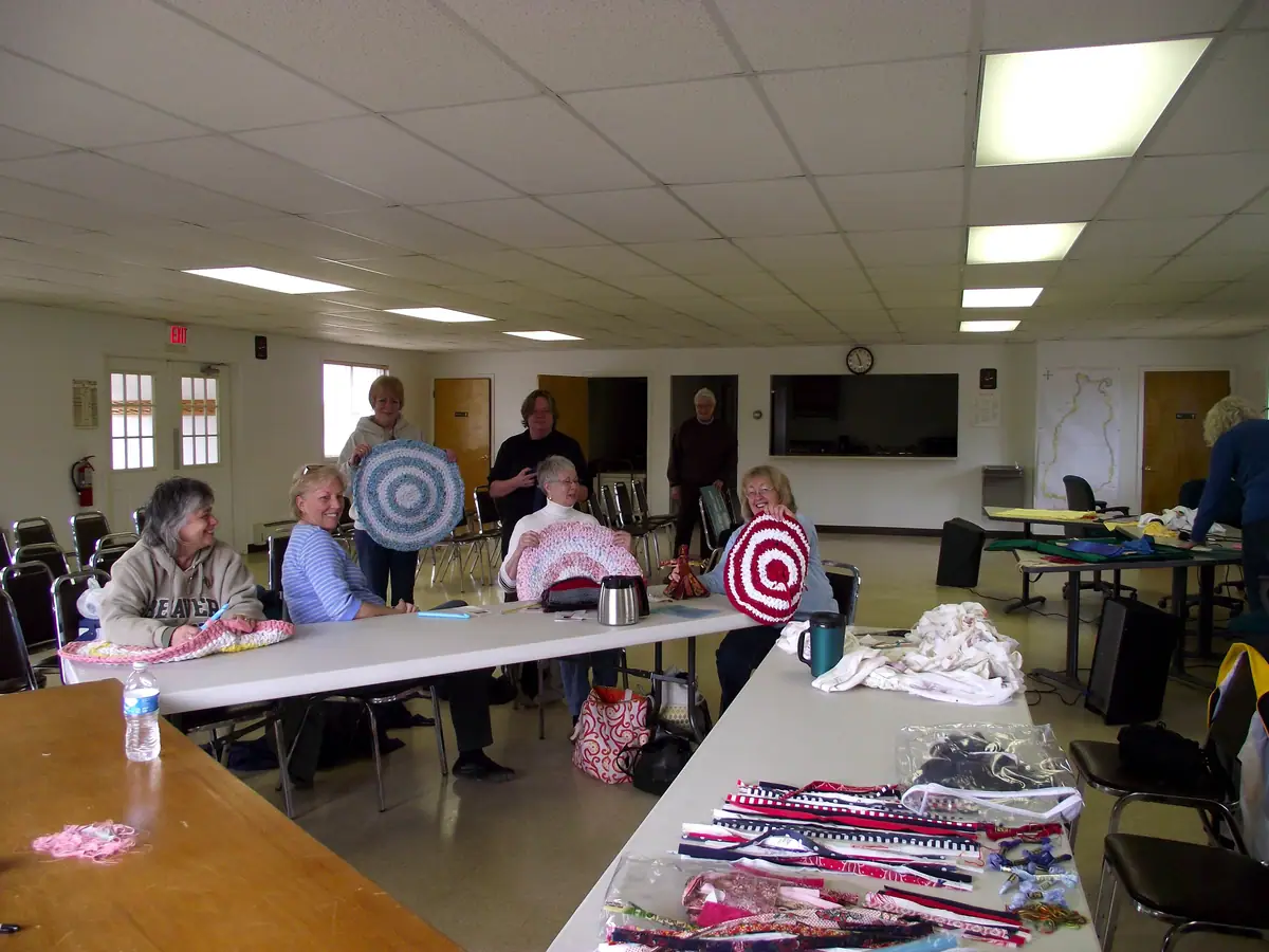 Women display round handmade rugs around tables in a bright room filled with textile craft materials.