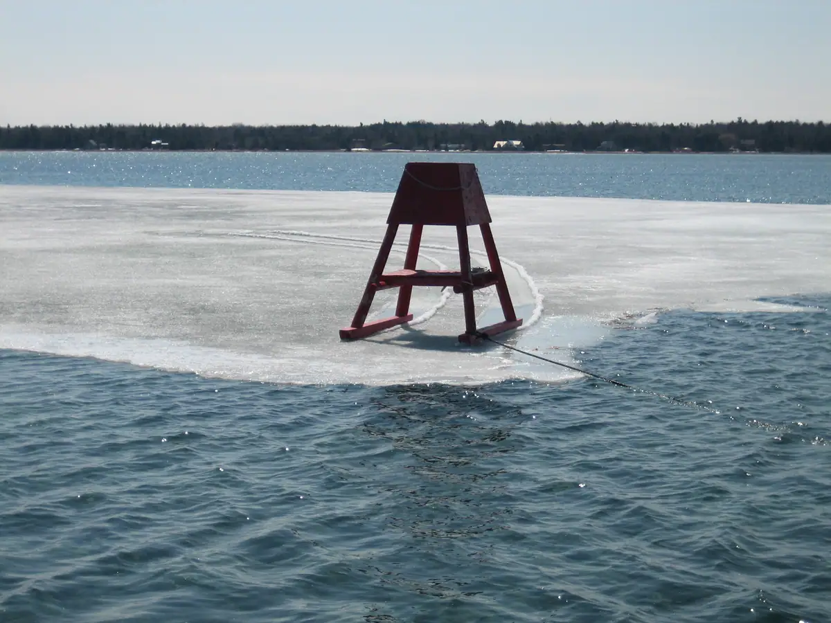A red A-frame structure stands on a sheet of ice beside open water, with a rope trailing into the foreground.
