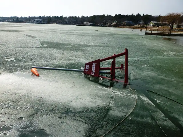 A red ICE CLASSIC marker lies partly submerged in cracked ice near a shoreline.
