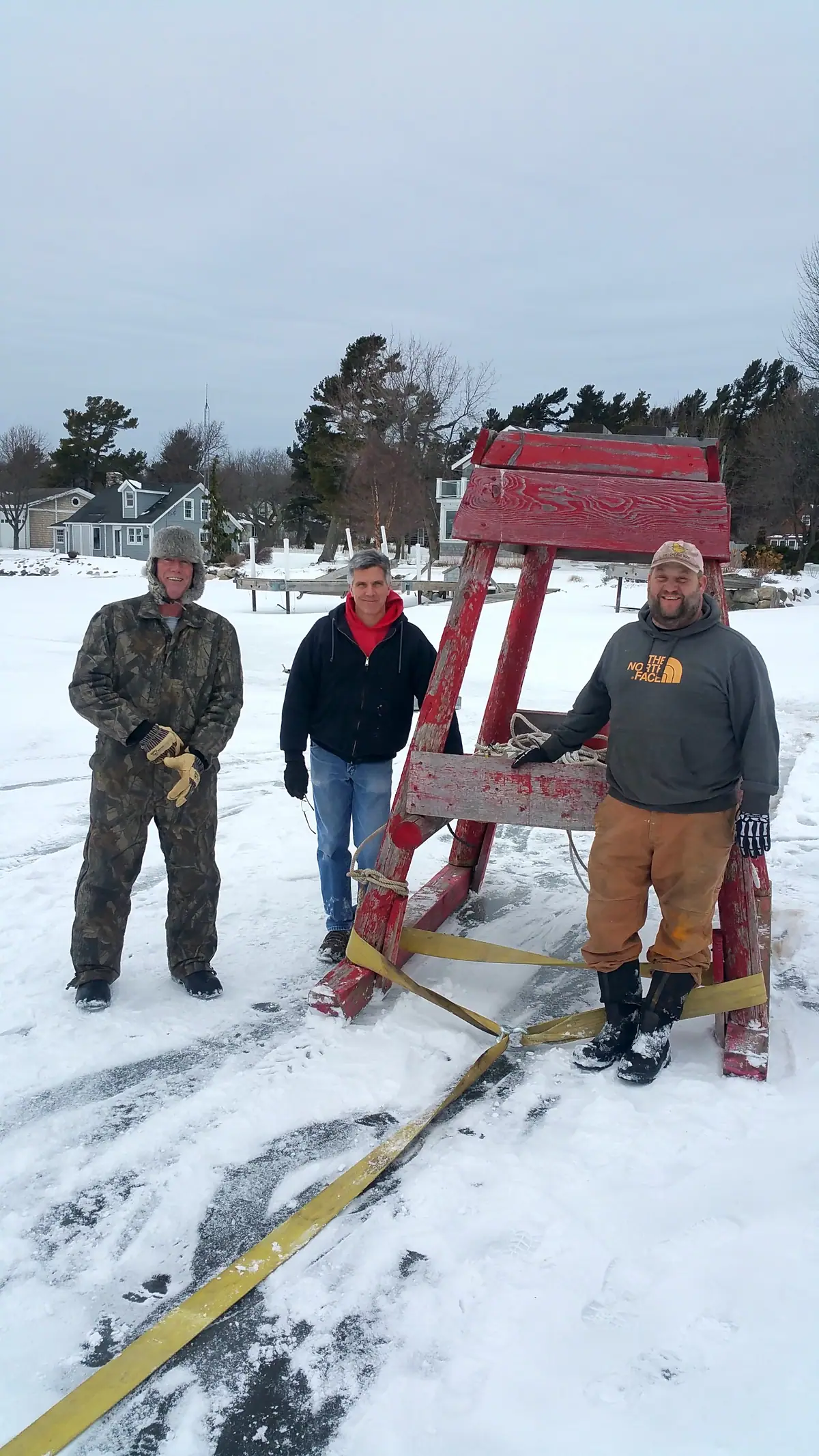 Three men stand beside a red wooden structure on a snow-covered frozen surface.
