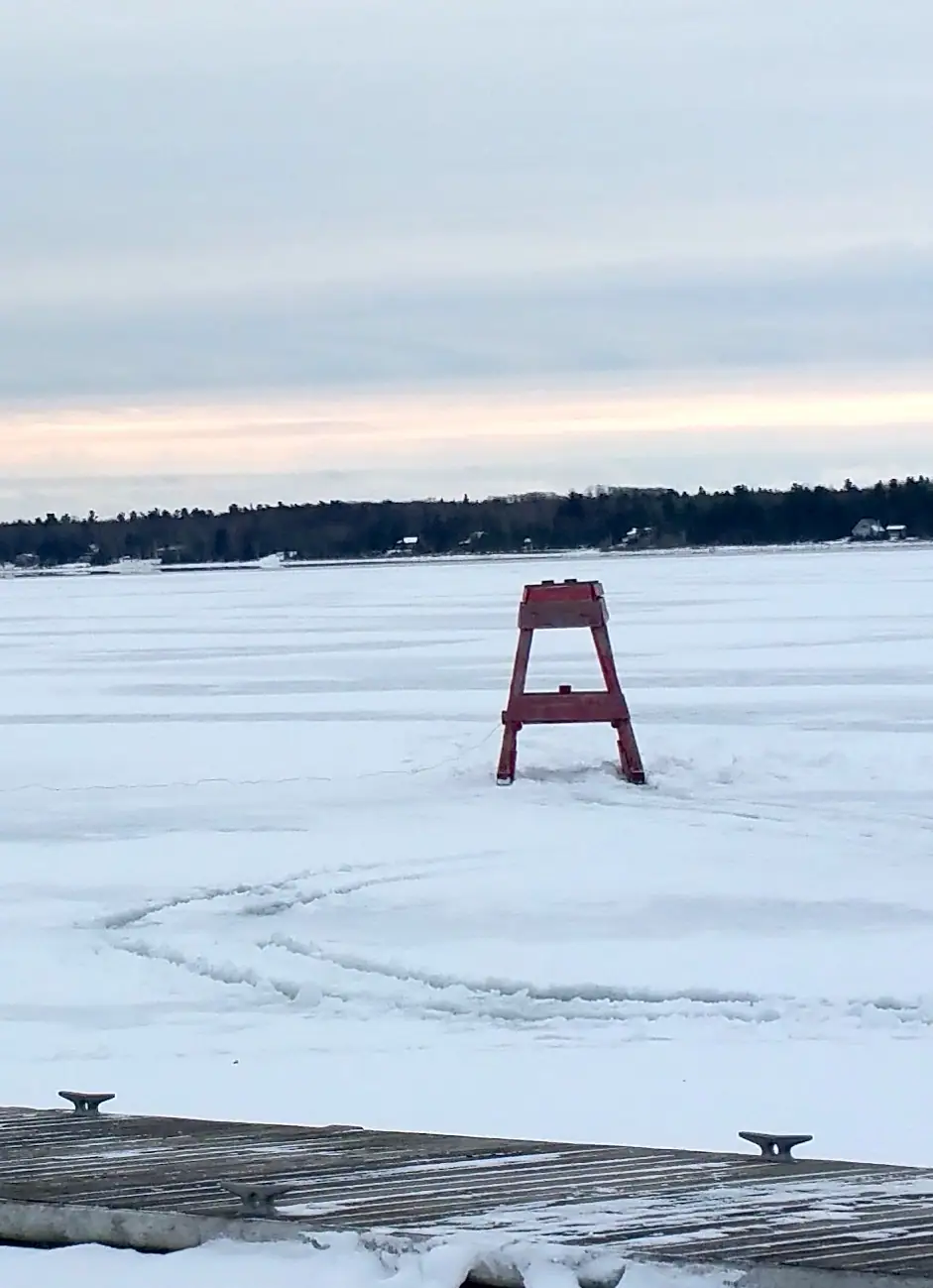 A red A-frame marker stands on frozen water beyond a snow-covered wooden dock.
