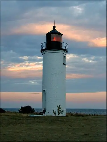 A white cylindrical lighthouse stands beside the shoreline beneath a pastel evening sky.