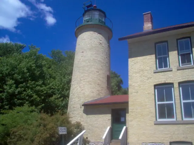 A pale brick lighthouse tower and adjoining building stand beneath a bright blue sky.