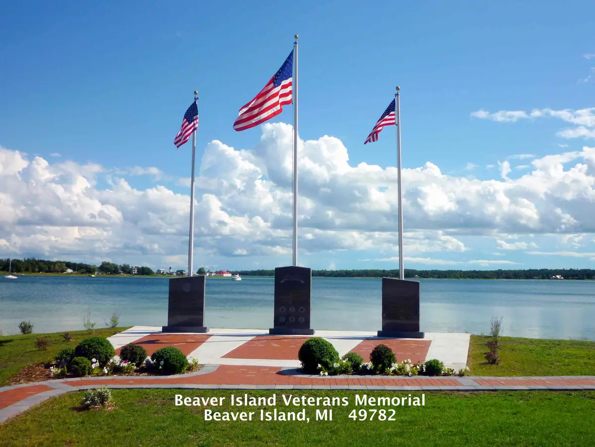 Three stone memorials and American flags stand on a landscaped waterfront plaza beneath a bright, cloud-filled sky.