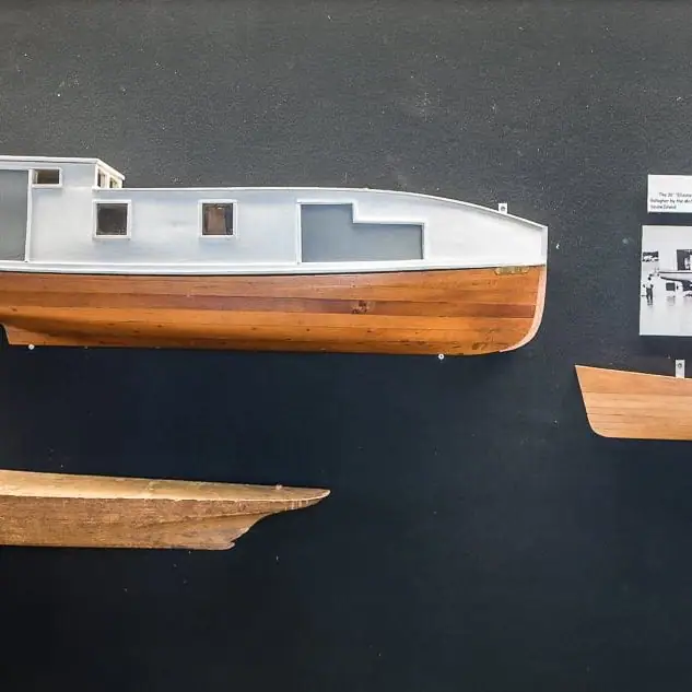 A wooden boat with a pale blue cabin is displayed against a dark wall beside partial wooden hulls.