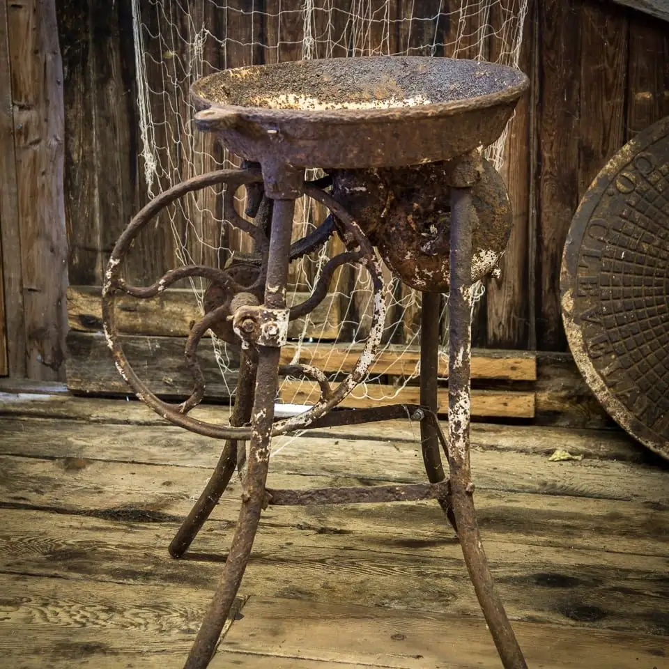 Rusty metal apparatus with a large wheel and shallow bowl stands on a weathered wooden floor.