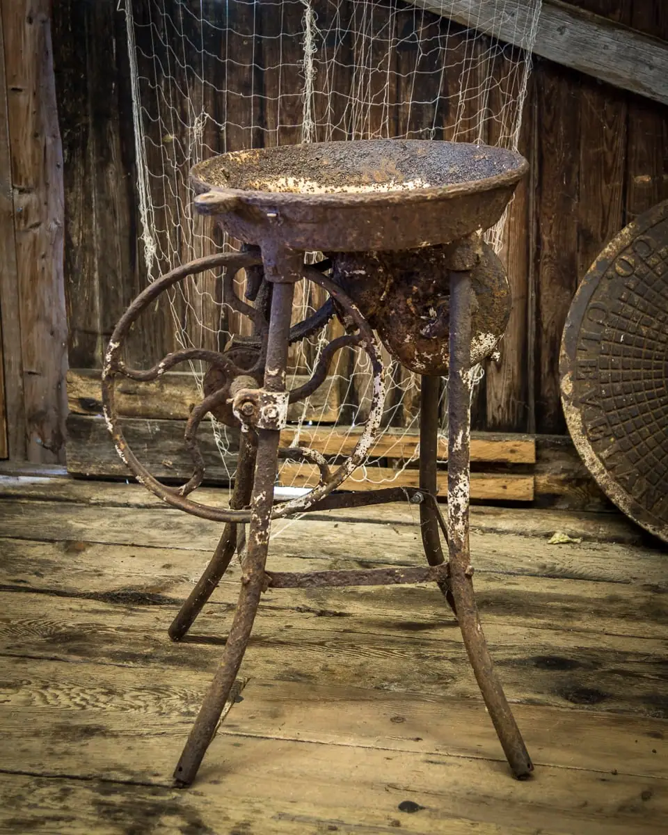 A heavily rusted metal apparatus with a shallow bowl, curved wheel-like supports, and legs stands on a wooden floor.