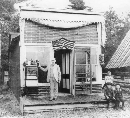 A man and three children stand outside a small brick storefront with a porch and display window.