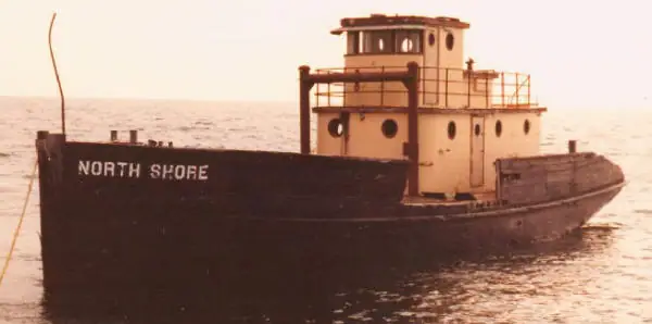 A tugboat named NORTH SHORE floats on calm water beneath a pale sky.