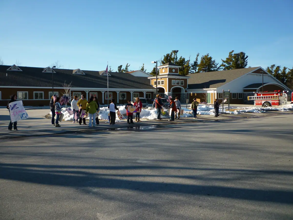 Children and adults hold handmade signs and flags outside a building beside snowbanks and a fire truck.