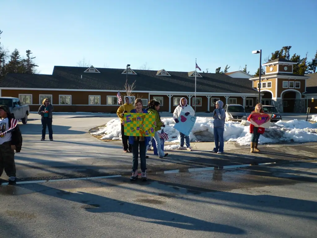 People holding colorful handmade signs in a snowy parking lot outside a long brown building.