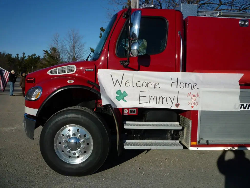 A red fire truck displays a handwritten “Welcome Home Emmy!” banner beside a roadside gathering.