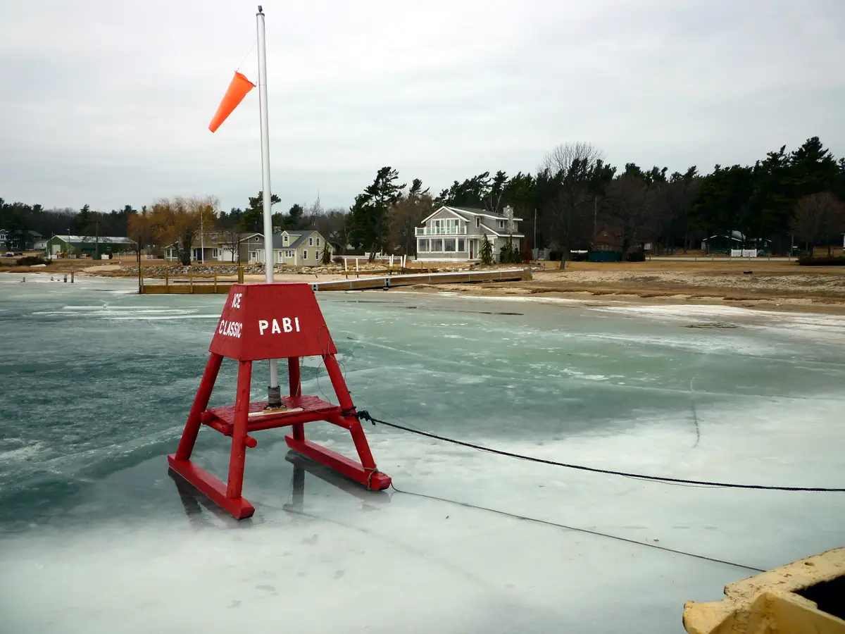 Red ice marker with an orange windsock stands on frozen water beside homes and a dock.