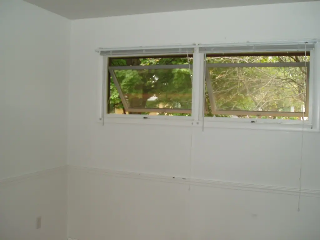 Empty white-walled room with two open windows, blinds, wall molding, and an electrical outlet.