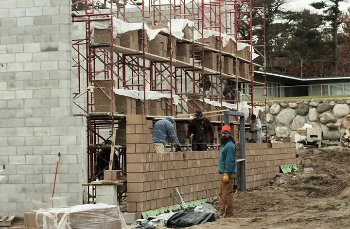 Workers build a concrete-block wall beside a scaffolded structure at a muddy construction site.