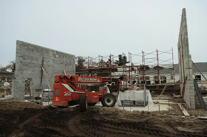 A red telehandler stands before partially built concrete-block walls and scaffolding at a muddy construction site.