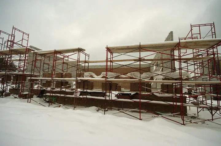 Snow-covered building construction site with red metal scaffolding and unfinished masonry walls.