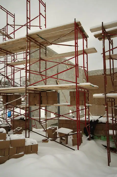 Snow-covered construction site with red scaffolding, unfinished masonry walls, lumber, and stacked blocks.