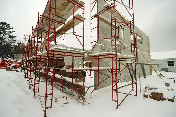 Snow-covered construction site with red scaffolding surrounding a concrete-block building.
