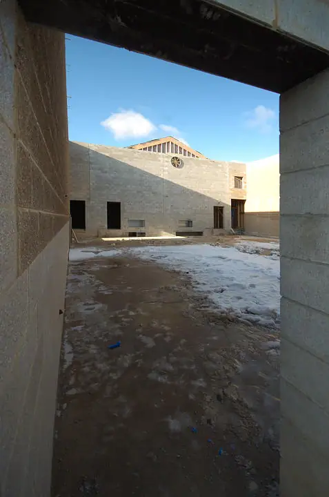 A snow-dusted construction courtyard is viewed through a concrete-block opening toward a partly built structure.