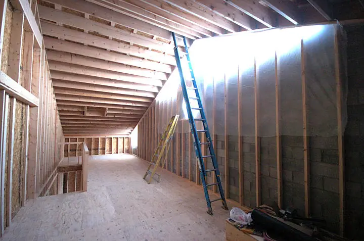 Unfinished interior with exposed wood framing, plywood flooring, plastic sheeting, and two ladders.