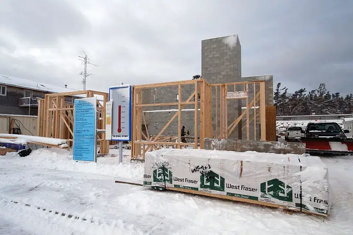 Snow surrounds a timber-framed building under construction, with wrapped lumber, a signboard, and parked trucks.