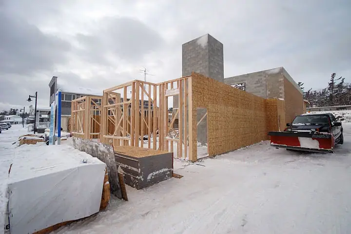 A timber-framed building rises beside a block chimney at a snow-covered construction site.