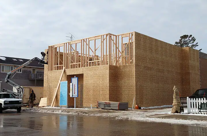 Wood-framed building under construction beside a wet, snow-lined street, with a lift and worker at the site.