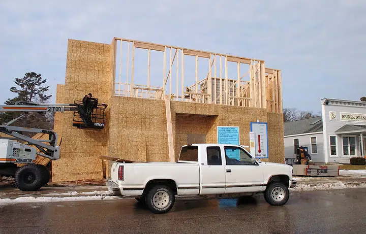 A wood-framed building under construction beside a wet street, with a pickup truck and boom lift in front.