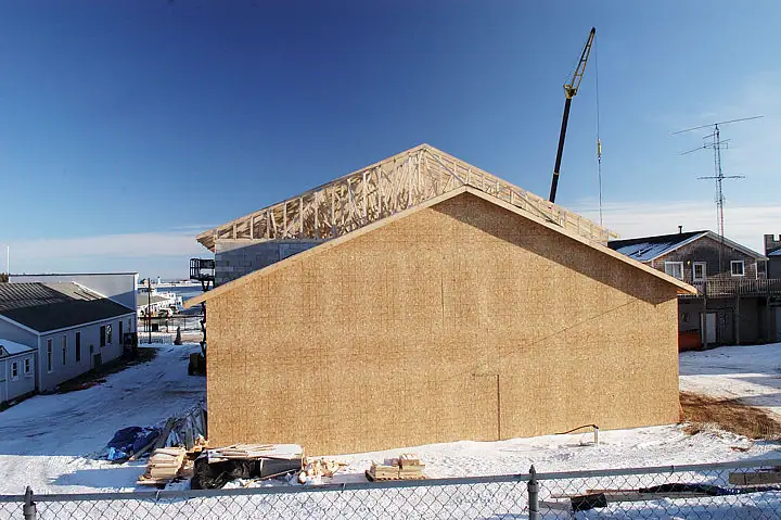 A sheathed building under construction with exposed roof trusses, a crane, snow, and a chain-link fence.