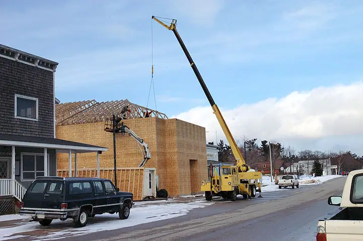 A crane lifts a roof truss onto a wooden building under construction beside a snowy street.