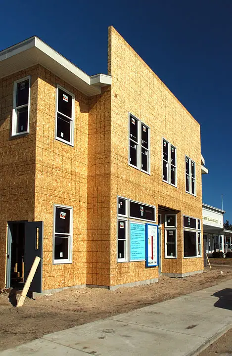 Two-story wood-framed building clad in OSB sheathing, with installed windows and a blue construction-code sign.