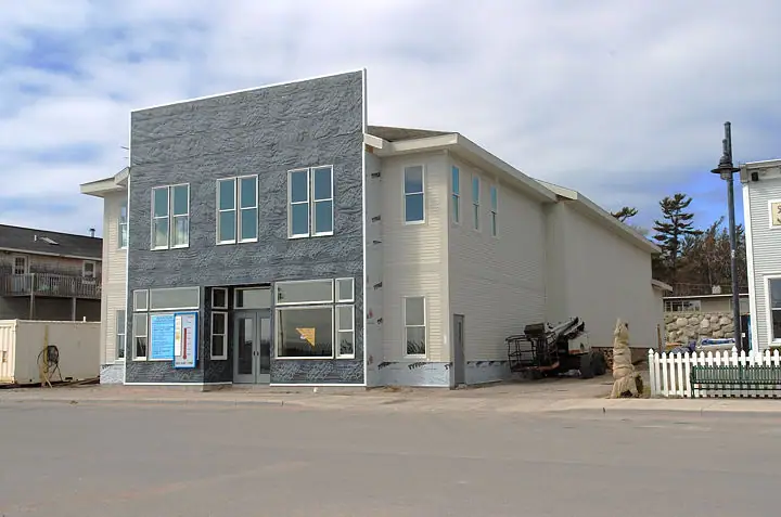 Two-story building with dark stone-textured facade, construction equipment, posters, and a white picket fence.