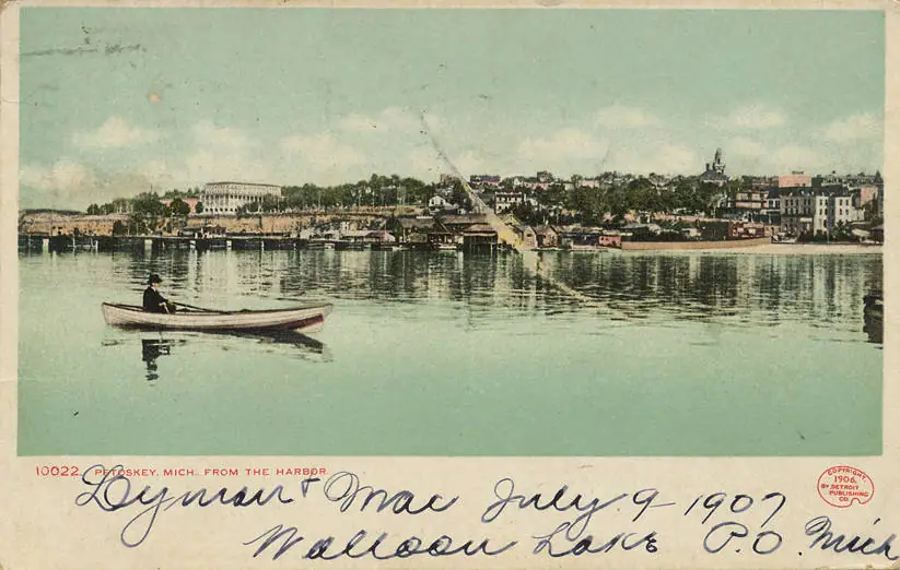 Colour postcard view across Little Traverse Bay to the Petoskey waterfront, with a rower in the foreground, docks and warehouses along the shore and the town rising behind.