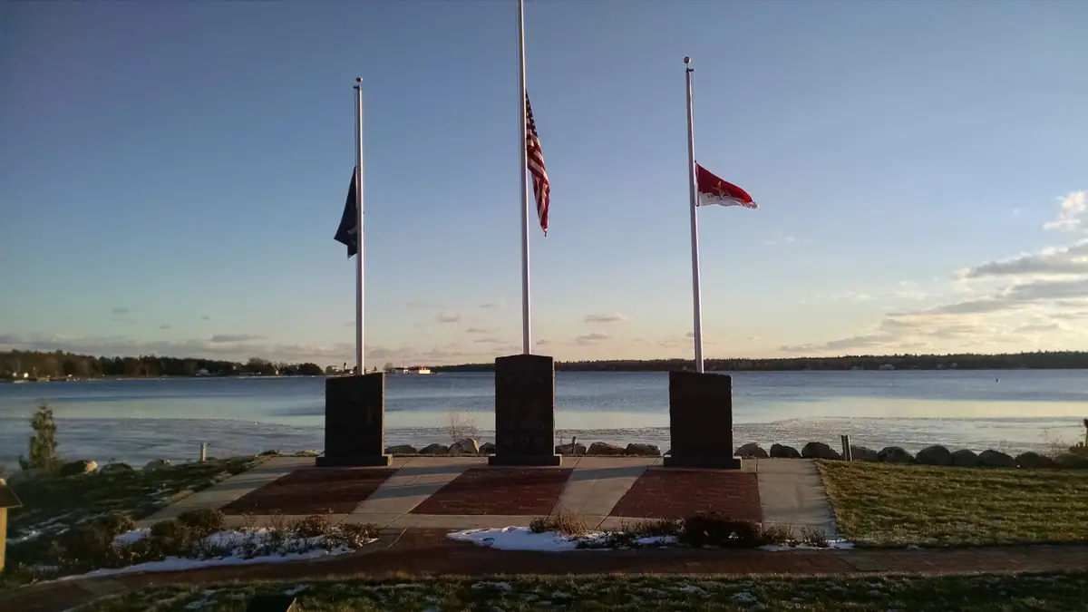 Three stone memorials and flagpoles stand beside the water in a paved plaza with patches of snow.