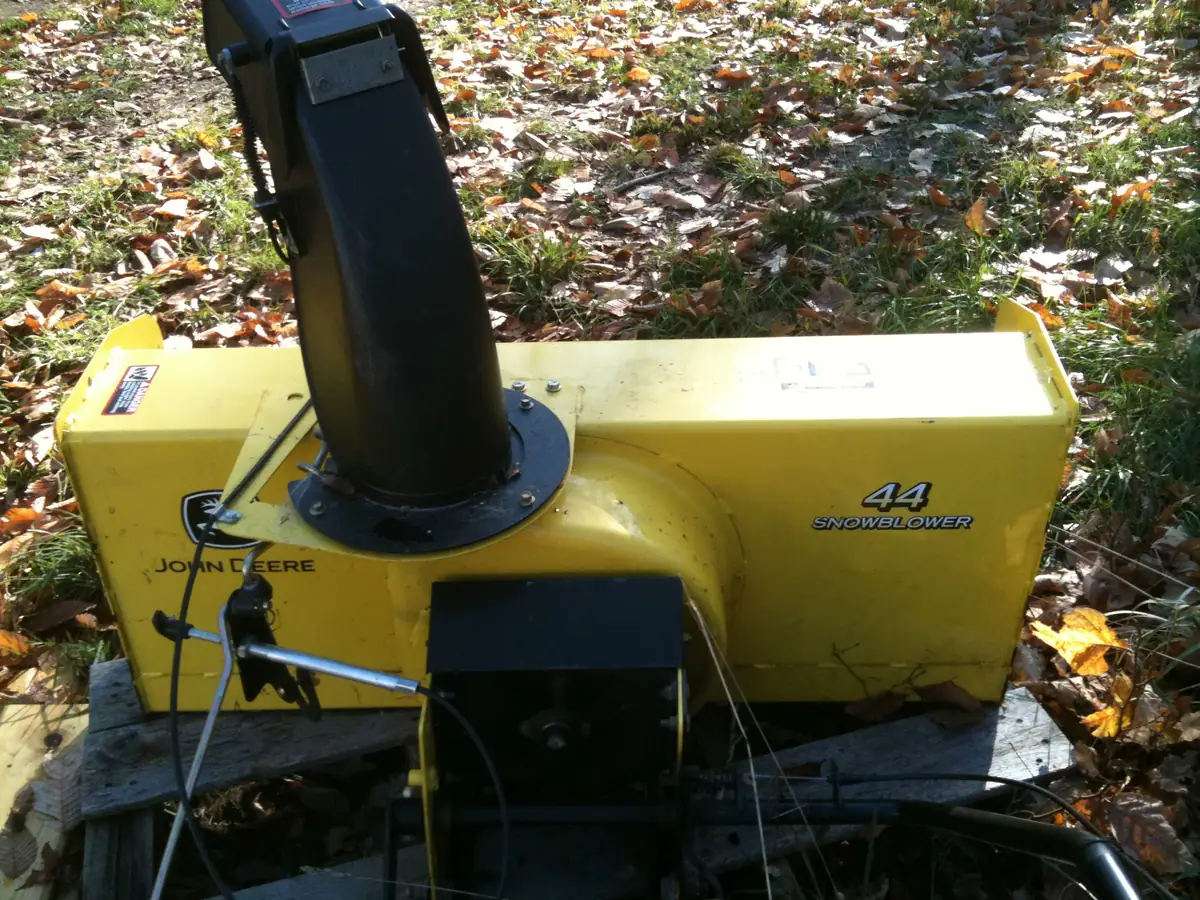 Yellow John Deere snowblower attachment resting on wooden planks beside leaf-covered grass.