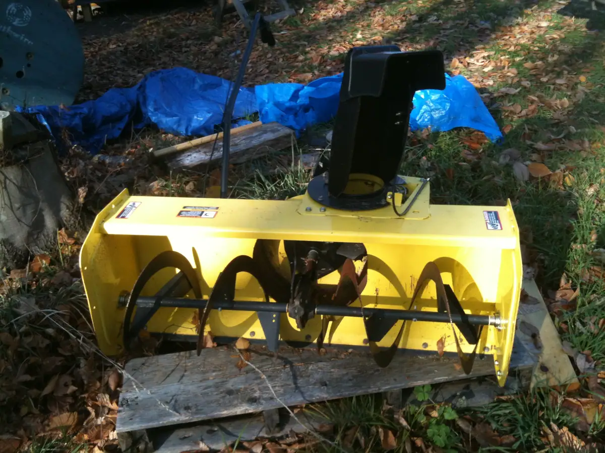Yellow auger-equipped outdoor machine resting on a weathered wooden board among fallen leaves.