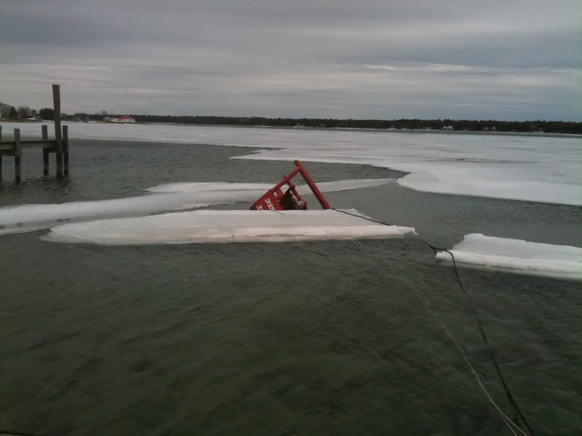 Broken ice surrounds a partly submerged red triangular frame near a wooden dock.